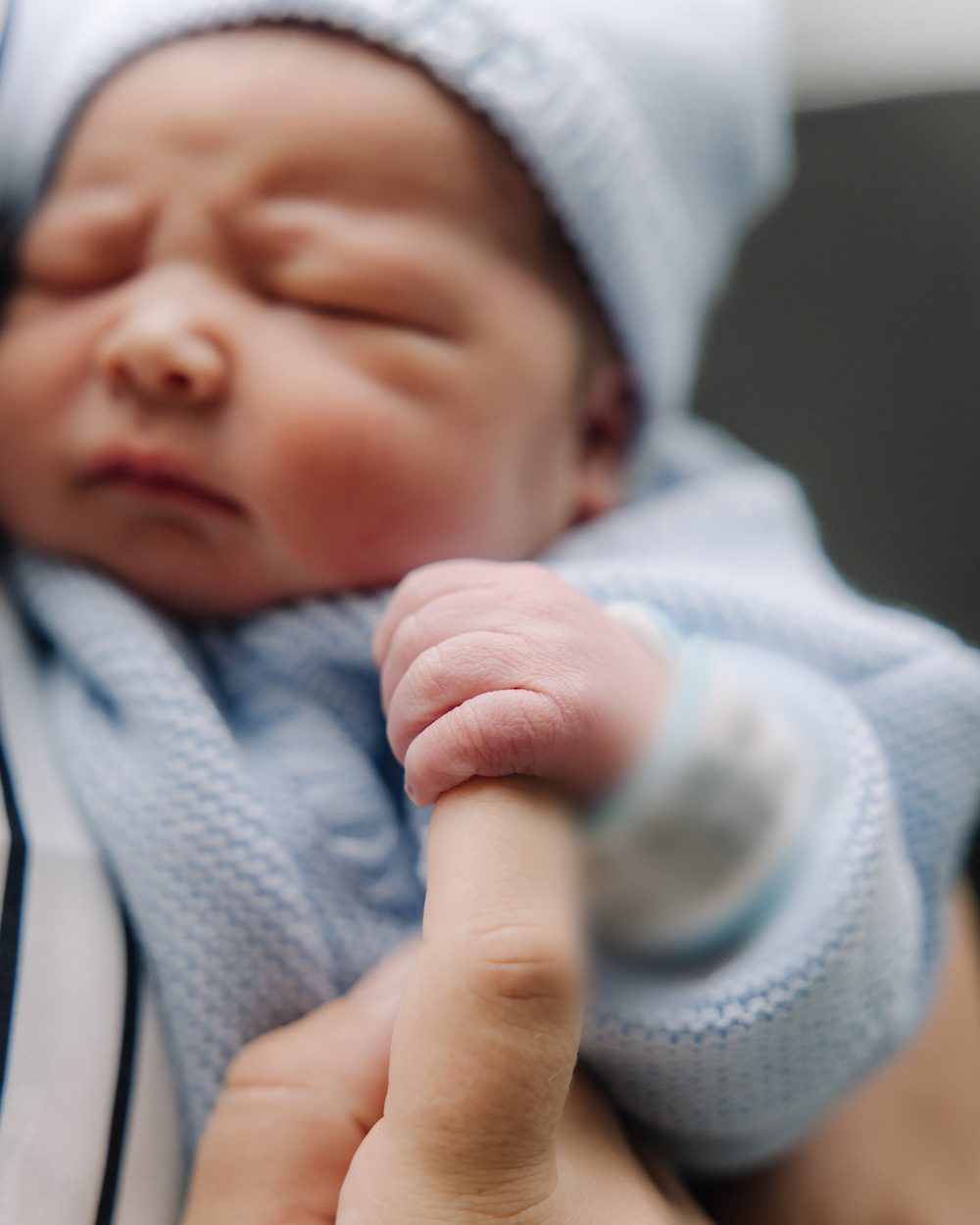 A newborn in a hospital cap gripping a parent's finger, hospital band on his wrist