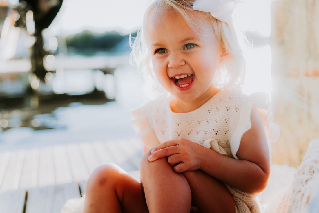 A toddler in a white knit dress and bow laughing on a sunlit dock