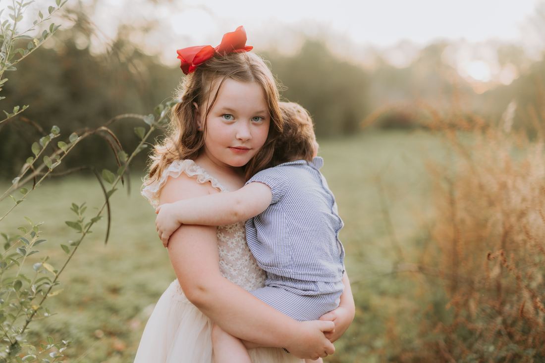 A girl in a red bow carrying her little brother on her hip in a golden field