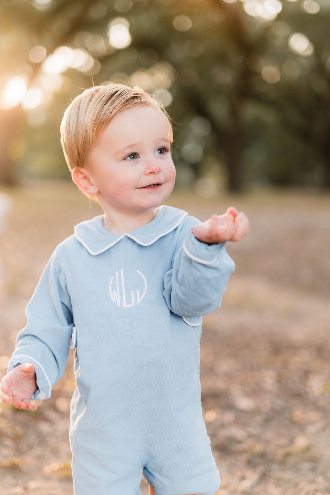 A toddler boy in a blue monogrammed romper reaching out in golden light