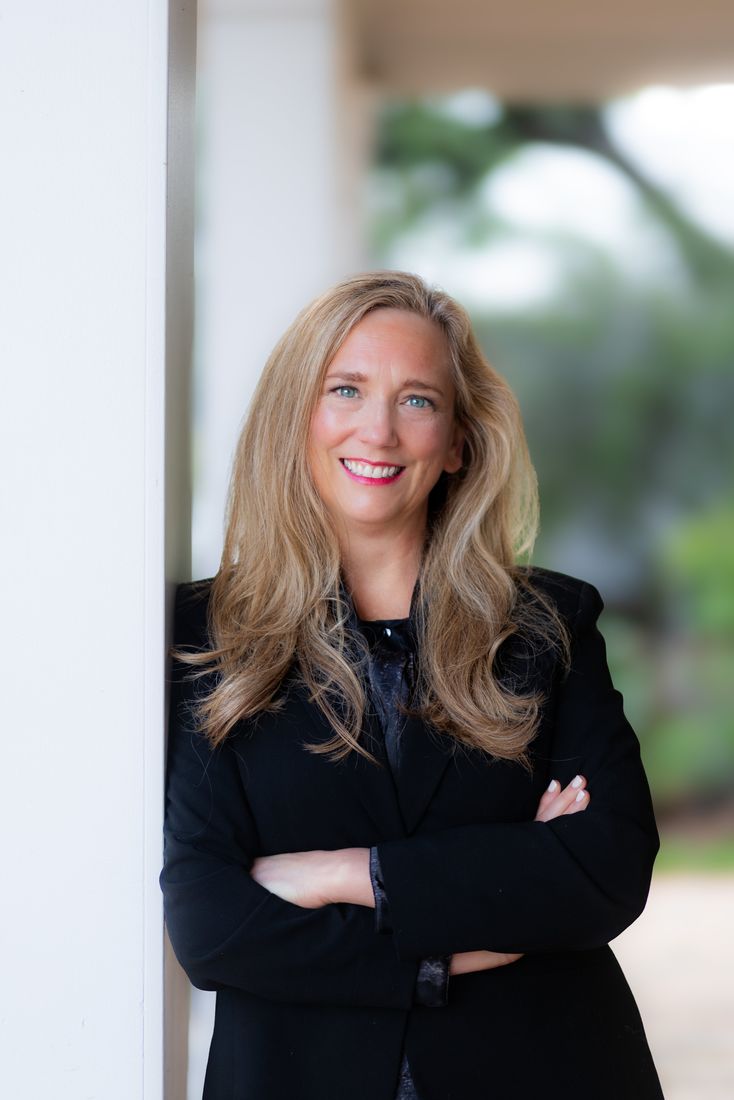 A woman in a black blazer leaning against a white column, arms crossed and smiling