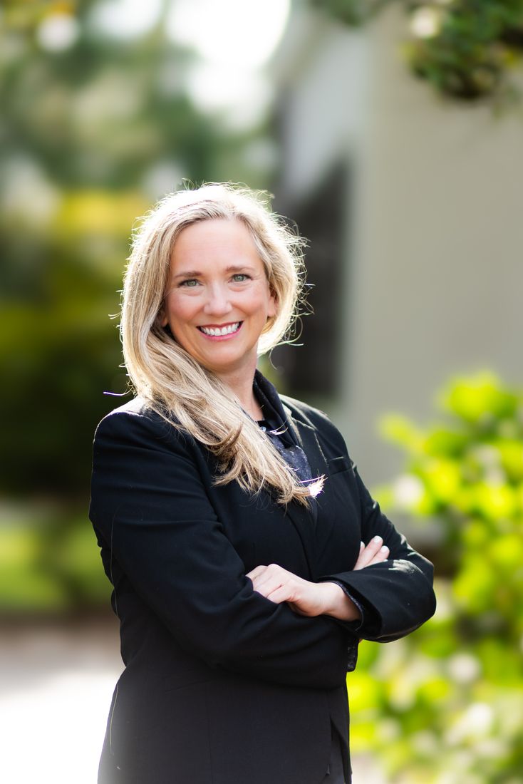A woman in a black blazer standing with her arms crossed, greenery soft behind her