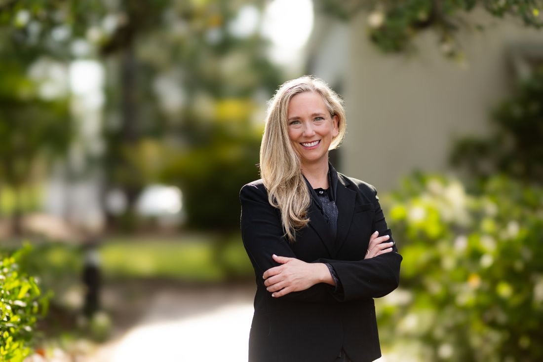 A wider headshot of a woman in a black blazer on a garden path, arms crossed