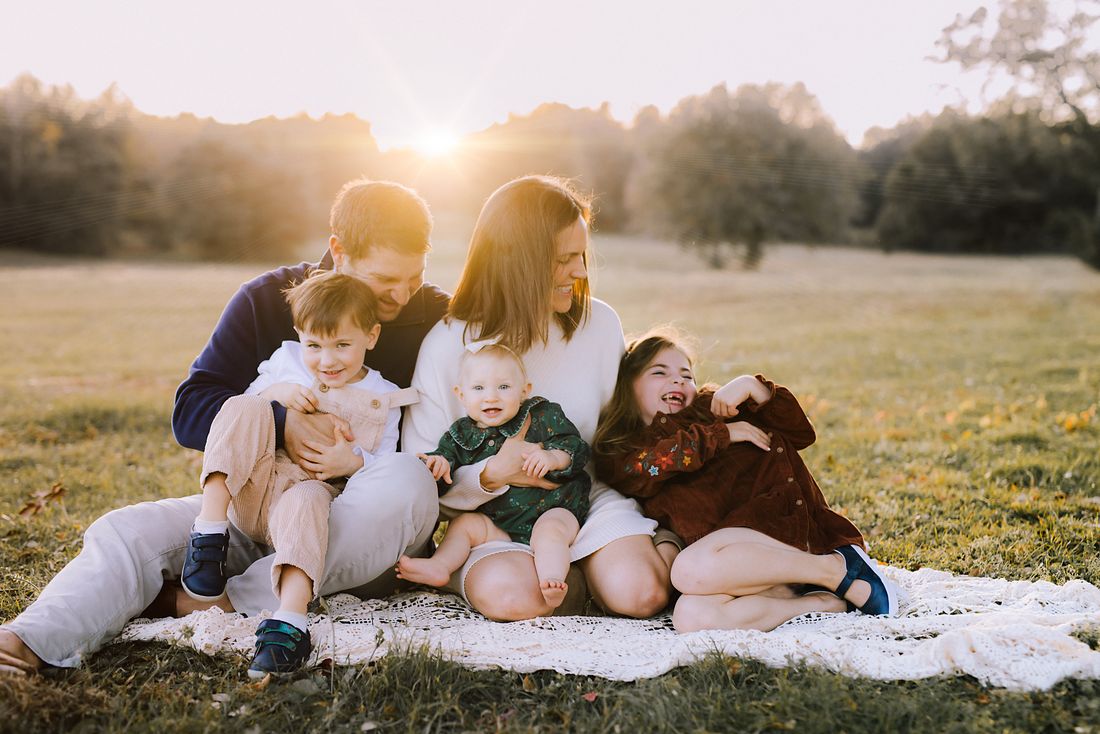 A family of five piled together on a lace blanket in a field, all laughing as the sun sets behind them