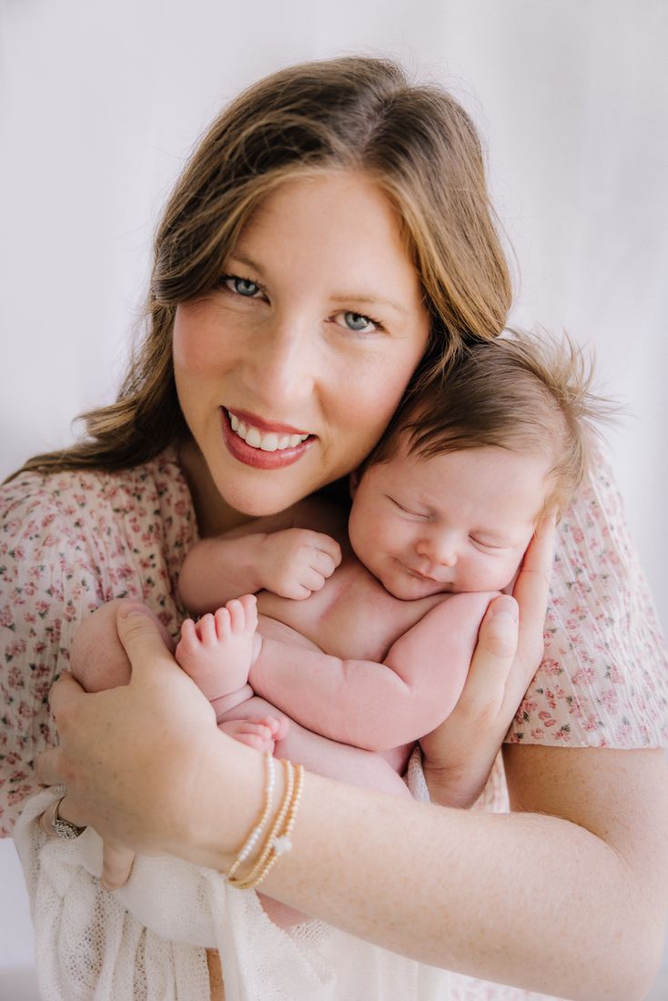 A mother in a floral dress smiling at the camera with her newborn curled against her chest