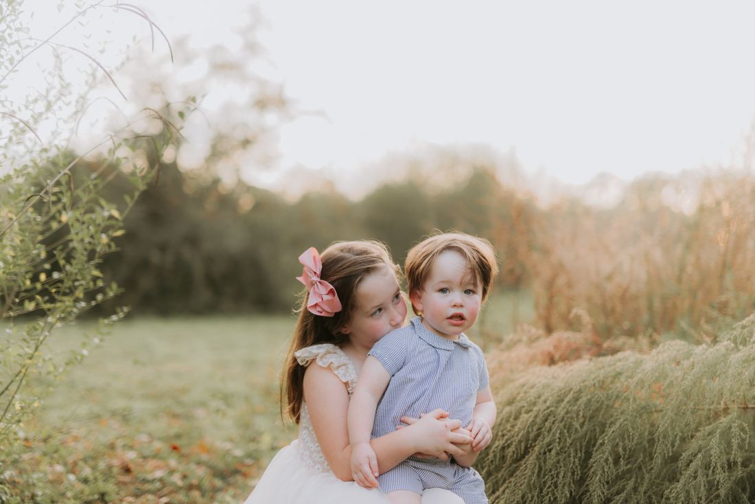 A big sister holding her baby brother on her lap in a field at sunset