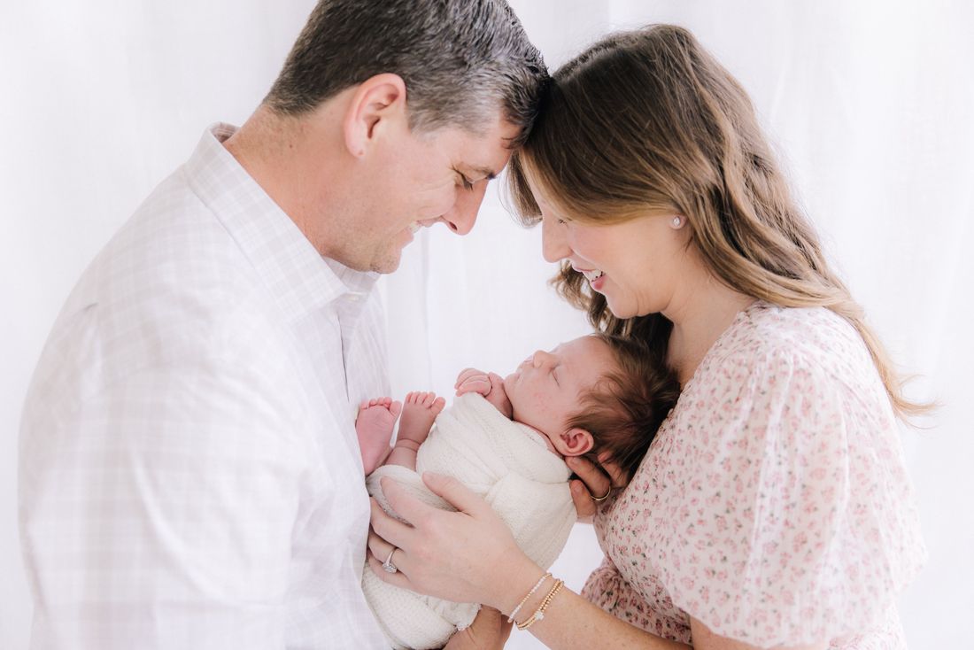 New parents forehead to forehead, smiling down at the swaddled newborn between them
