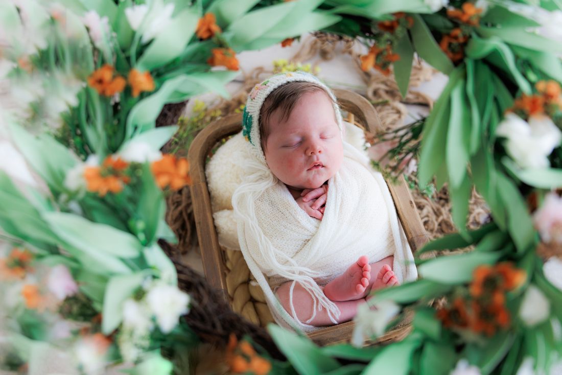 A newborn in a knit bonnet asleep in a wooden crate, framed by orange and white blooms
