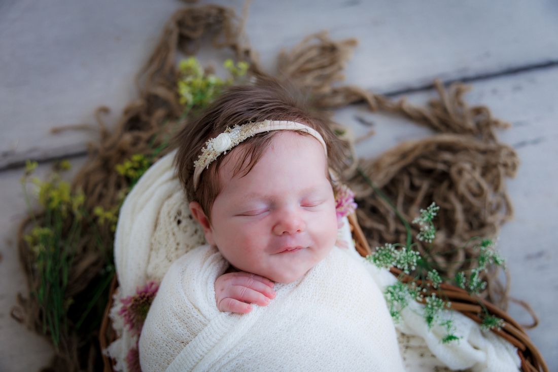 A swaddled newborn asleep in a wooden crate with greenery and rope around her