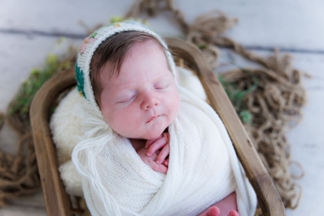 Close view of a swaddled newborn in a knit bonnet, hands tucked up under her chin
