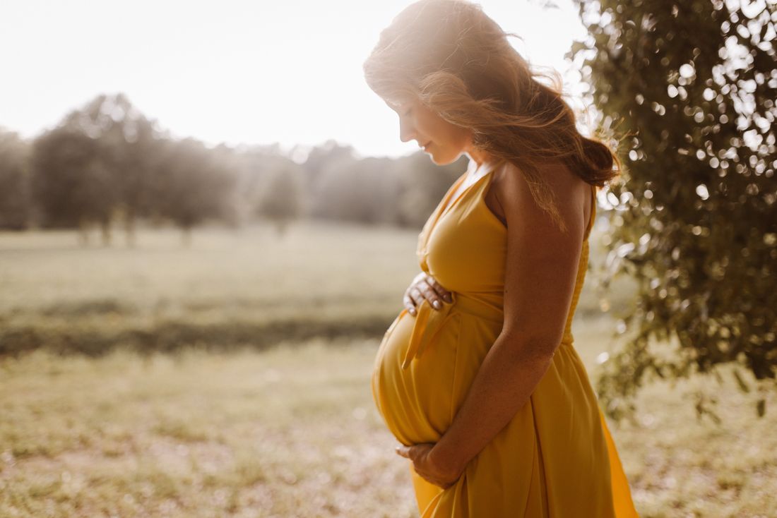 An expecting mother in a golden yellow dress looking down at her belly in a sunlit field