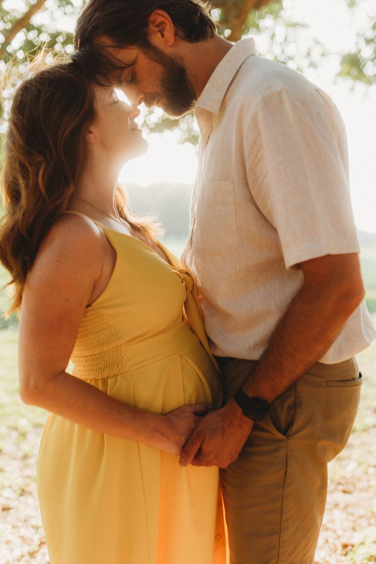 An expecting couple standing forehead to forehead, both hands resting on her belly in the backlight