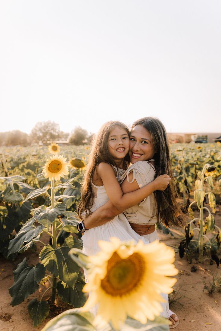 A mother holding her daughter on her hip in a sunflower field, both smiling at the camera