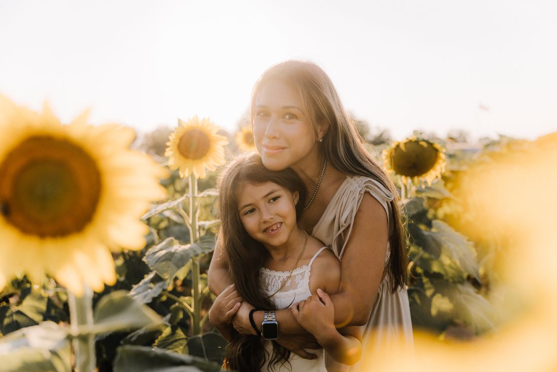 A mother with her arms wrapped around her daughter among the sunflowers at golden hour