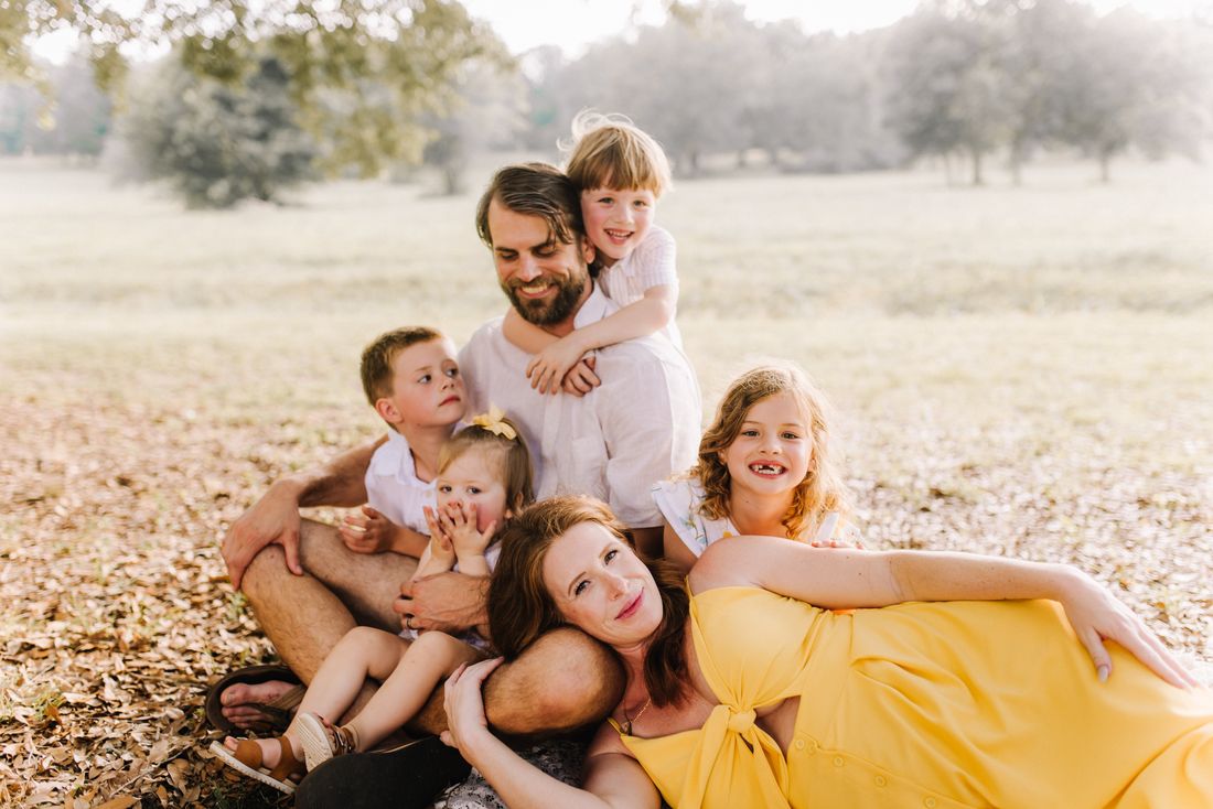 A family of six leaning on each other in a field of fallen leaves, the mother in yellow resting on her husband's knee