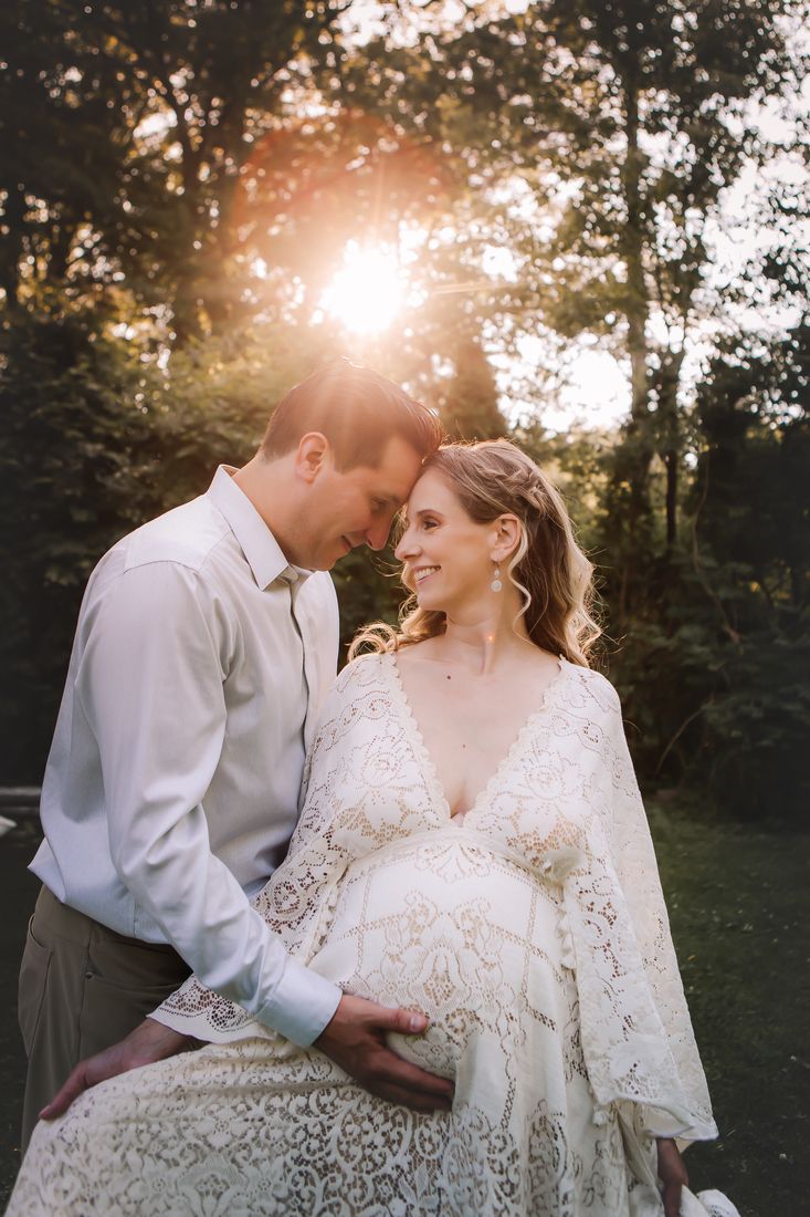 An expecting couple leaning their foreheads together in a cream lace gown, his hand resting on her belly in the backlight