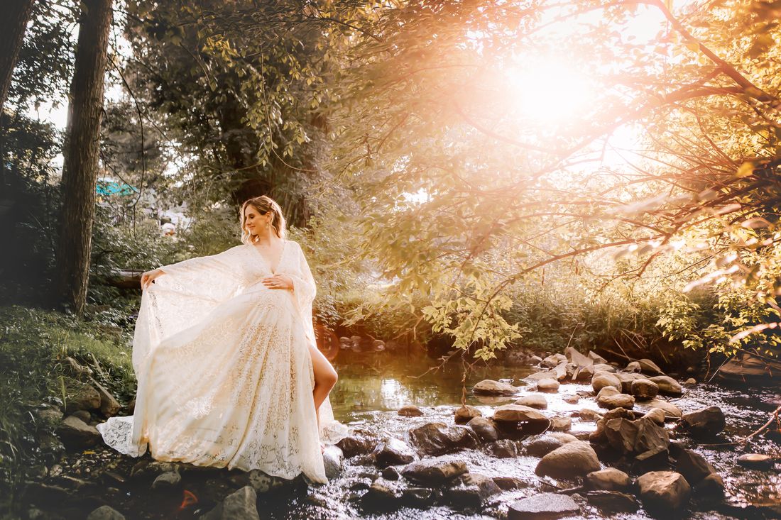 An expecting mother sweeping the train of her lace gown at the edge of a sunlit creek