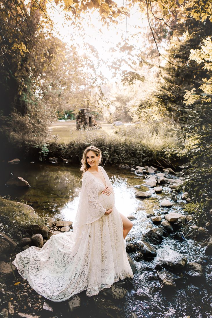 An expecting mother standing in a creek, her lace gown spread out across the rocks behind her