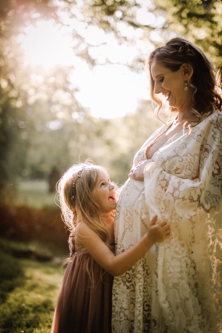 A little girl in a rose dress hugging her expecting mother's belly, the two of them laughing at each other