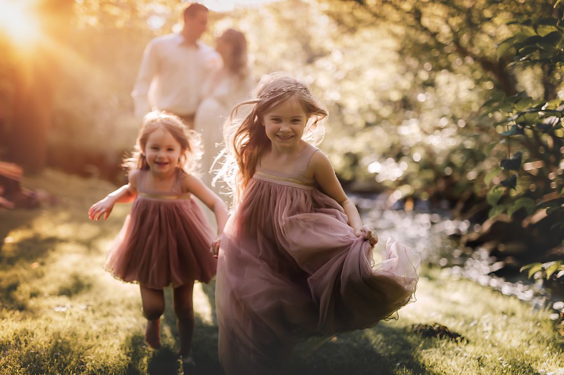 Two sisters in rose tulle running through the grass, their expecting parents watching from behind