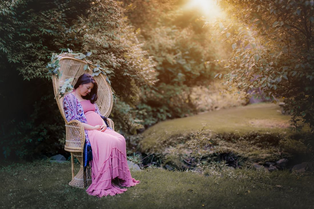 An expecting mother in a rose dress seated in a peacock chair at the edge of a garden, looking down at her belly