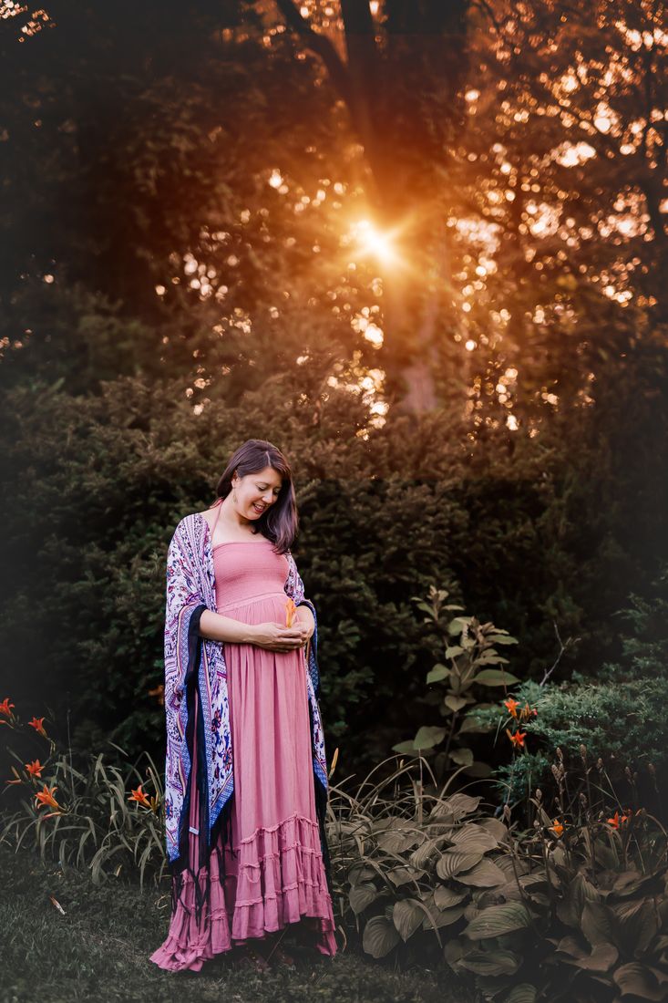 An expecting mother in a rose dress and patterned kimono standing among daylilies, the sun setting behind the trees