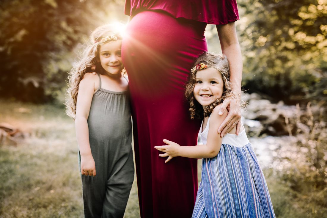 An expecting mother in a deep red gown beside a creek, one hand resting under her belly