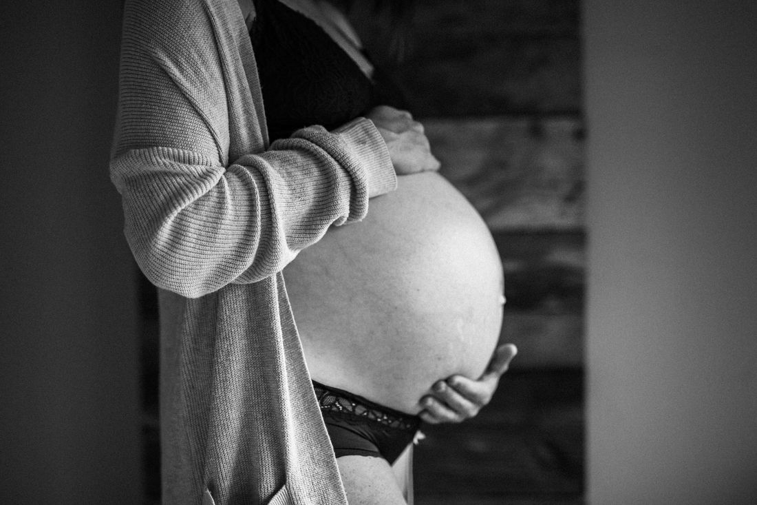 Black and white profile of an expecting mother in a knit cardigan, both hands beneath her belly in quiet indoor light