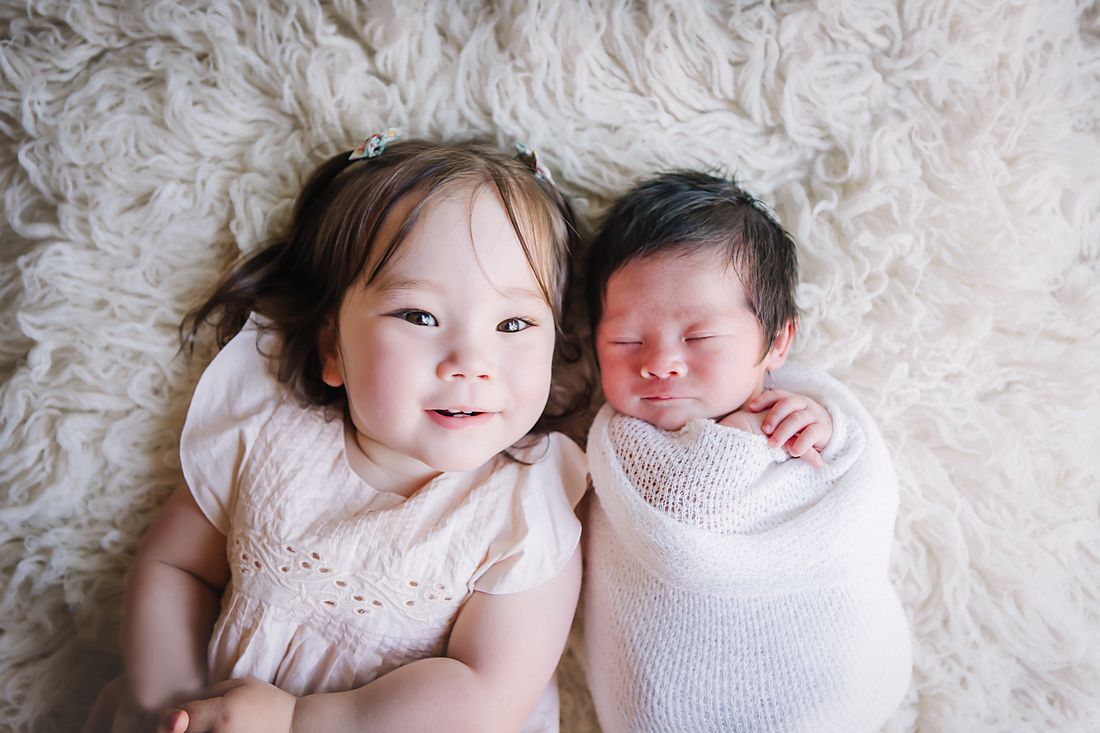 A toddler in a cream dress lying beside her swaddled newborn sibling on a flokati rug