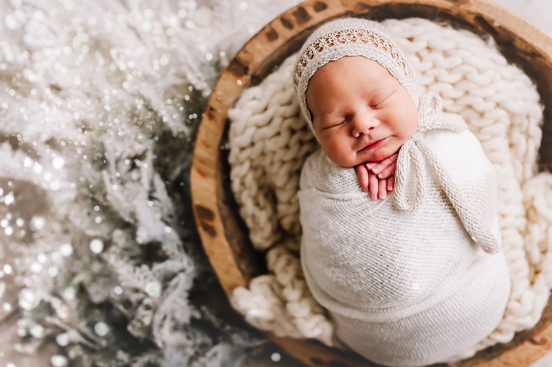 A newborn in a knit bonnet asleep in a wooden bowl, soft winter greenery beside her