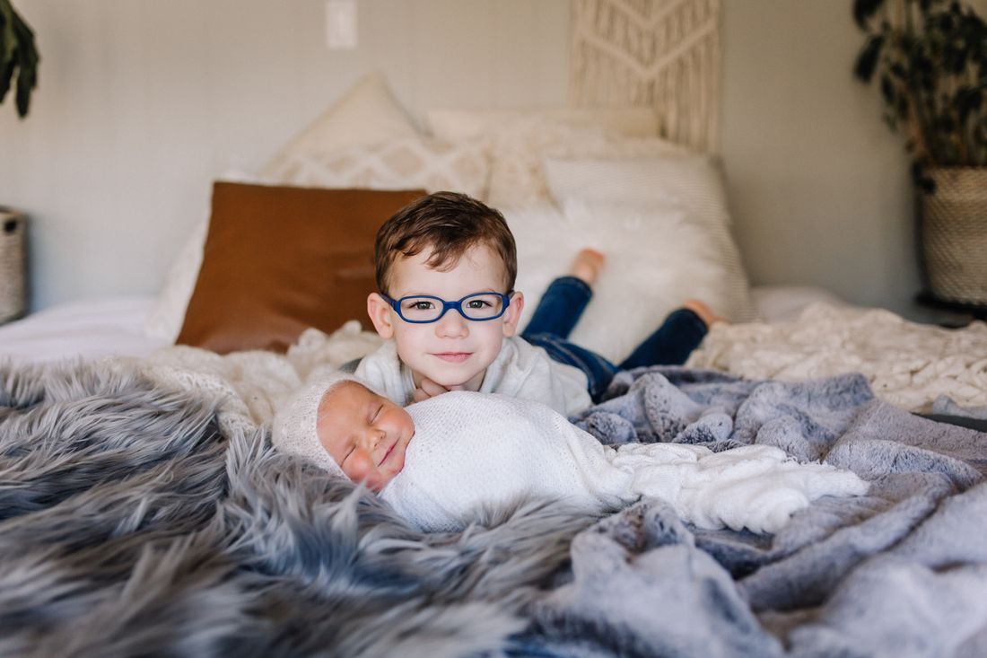 A big brother in blue glasses lying on the bed beside his swaddled newborn sibling