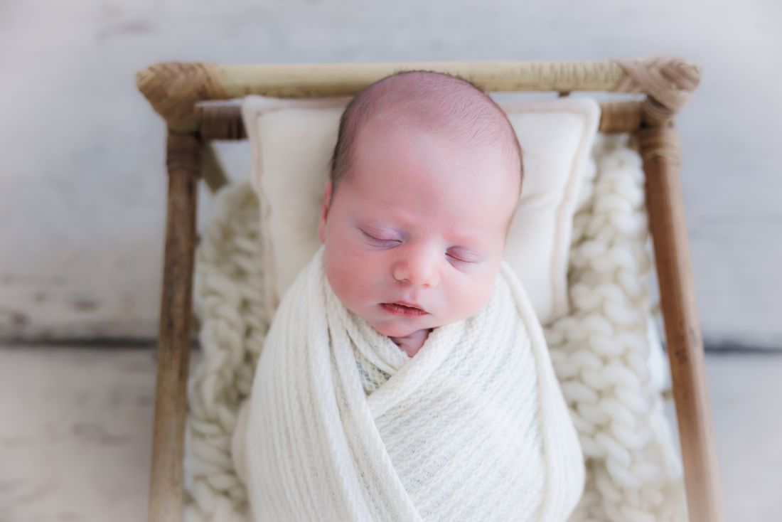 A swaddled newborn asleep on a small rattan bed against a chunky knit blanket