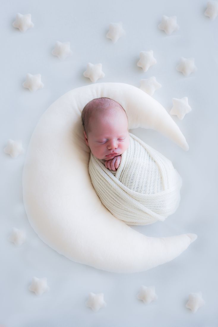 A swaddled newborn curled inside a crescent moon pillow, felt stars scattered around