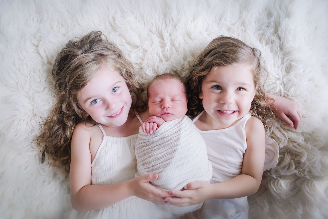 Two big sisters lying on either side of their swaddled newborn sibling, holding him between them