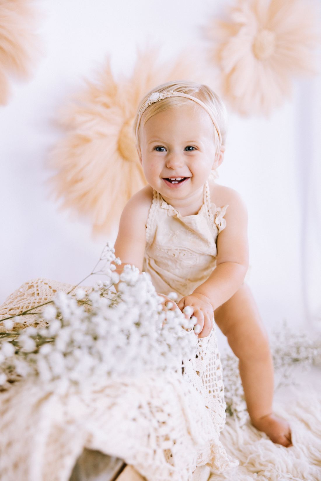 A baby girl in a lace romper and headband grinning beside a spray of baby's breath