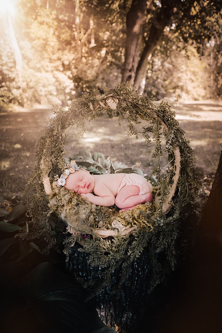 A newborn in a flower headband asleep inside a greenery-wrapped hoop hung between the trees