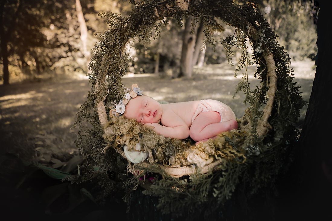 A newborn curled asleep on curly wool inside a greenery hoop, the woods soft behind her