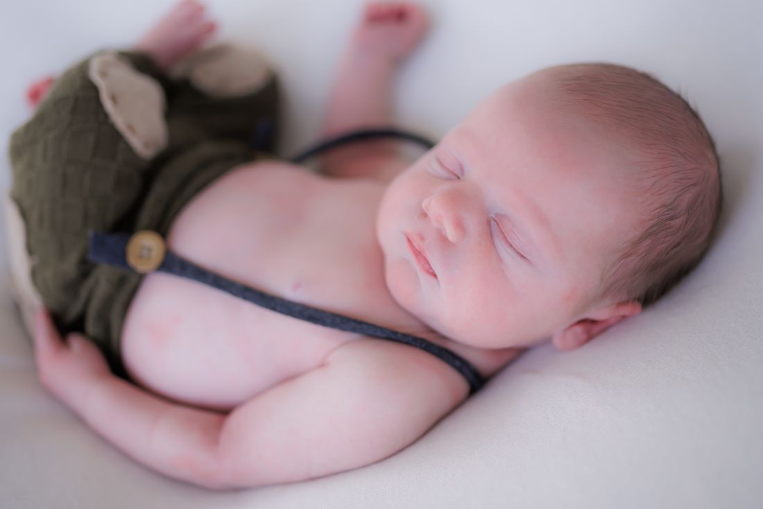A newborn asleep in an olive knit romper with suspenders and a matching bonnet