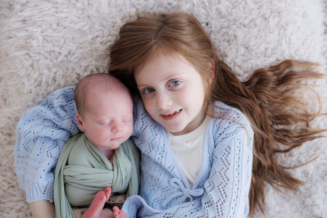 A big sister lying beside her newborn brother on a flokati rug, both in soft blue