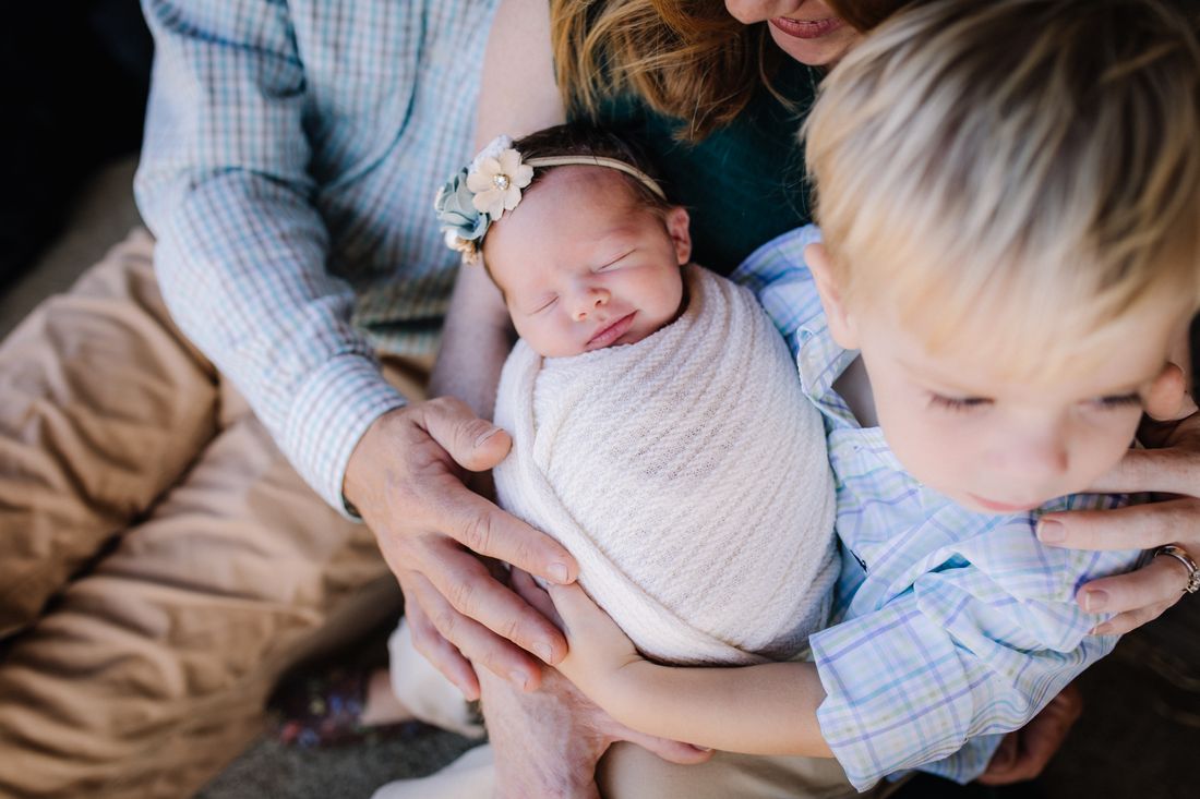 A newborn in a flower headband held in her family's hands, her big brother leaning in beside her