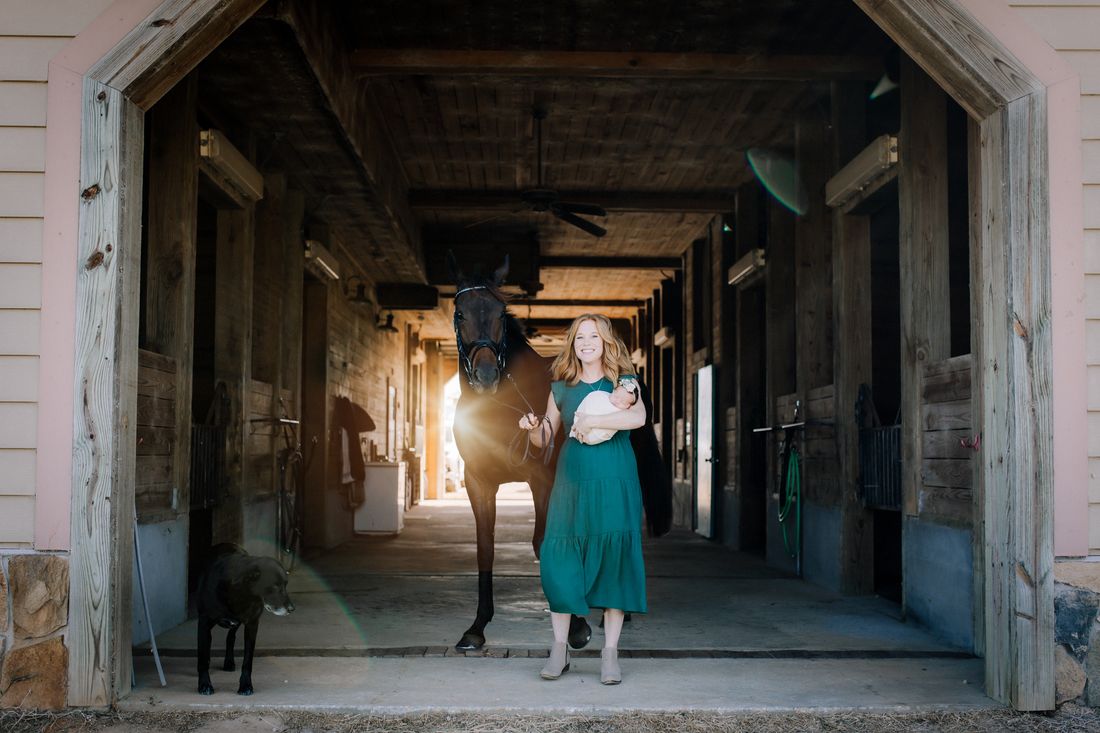 A mother in a green dress carrying her swaddled newborn down the barn aisle, leading her horse beside her