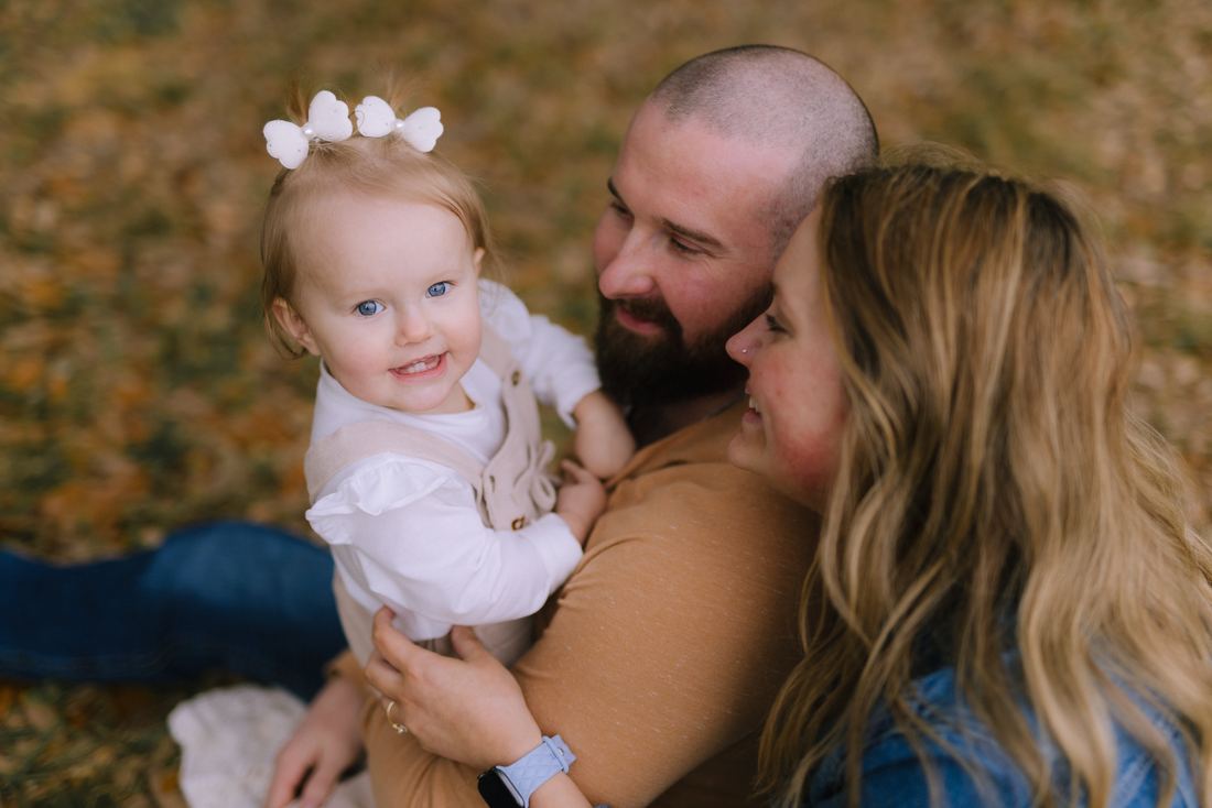 A blue-eyed toddler in a white bow smiling over her mother's shoulder while both parents look at her, the autumn leaves gold behind them