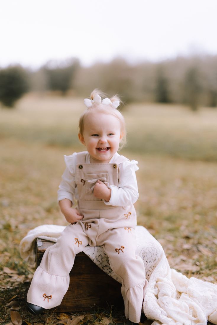 A toddler in bow-embroidered corduroy overalls sitting on a wooden crate in an open field, laughing wide at the camera