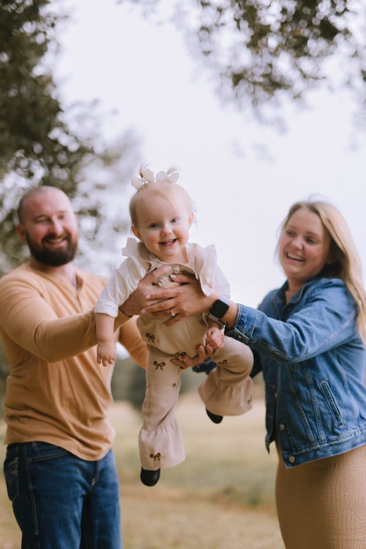 Two parents holding their laughing daughter up between them beneath a live oak, everything soft and bright behind