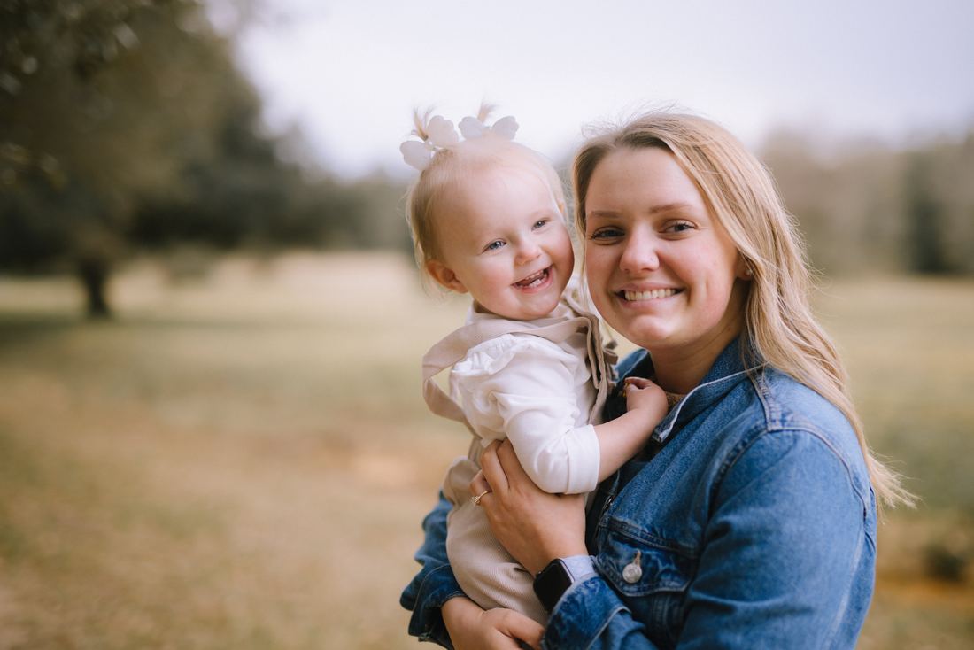 A mother in a denim jacket holding her toddler daughter cheek to cheek, both grinning at the camera in an open field