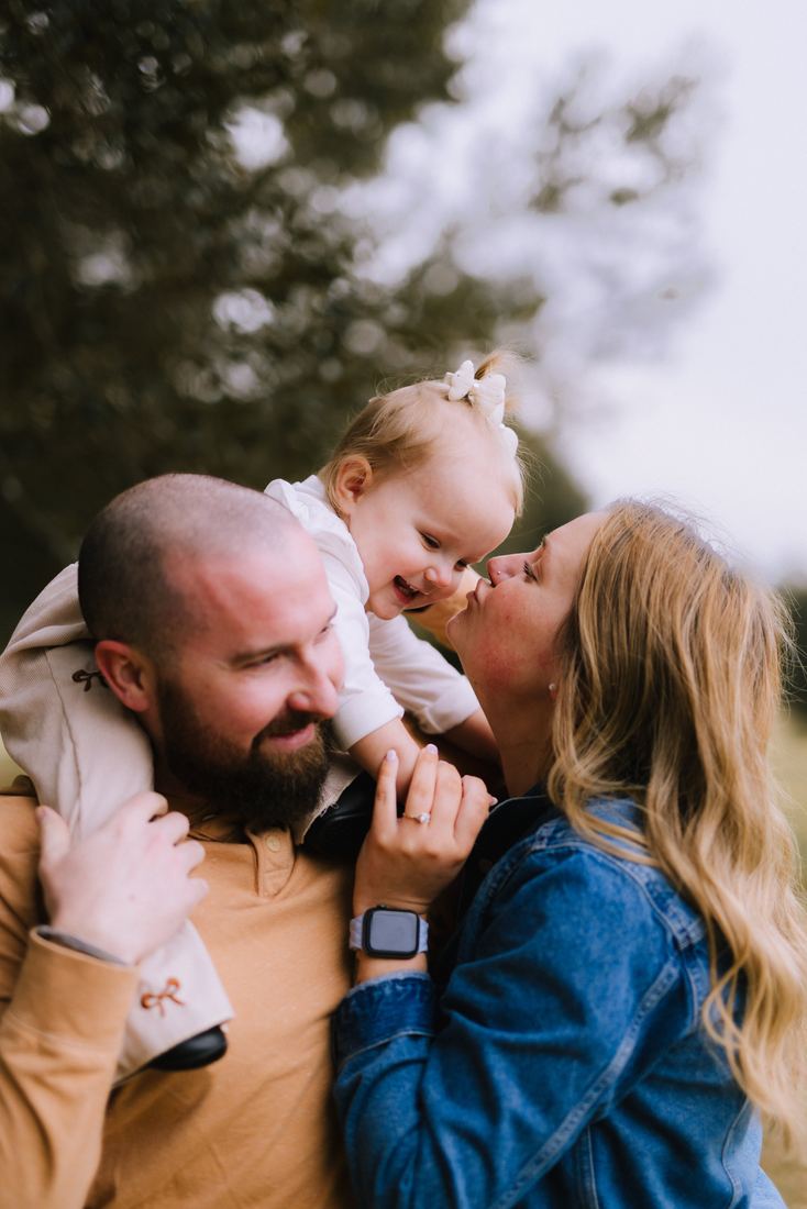 A little girl perched on her father's shoulder laughing while her mother leans in to kiss her cheek