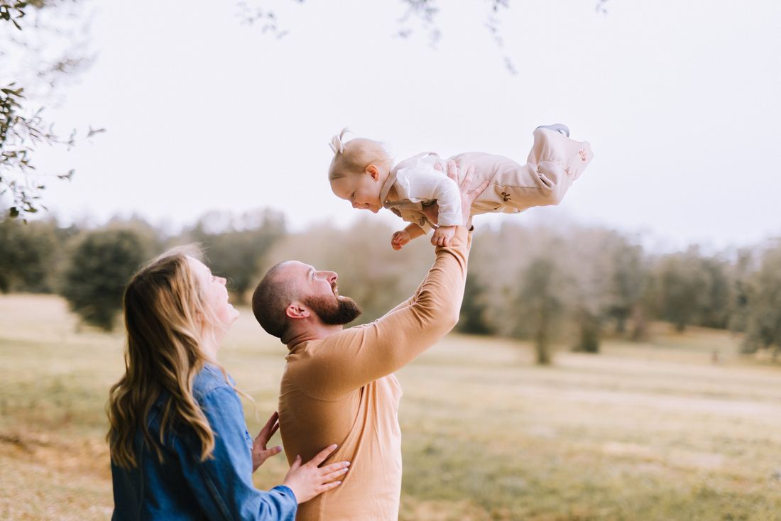 A father lifting his baby daughter overhead against a bright sky while her mother watches and laughs