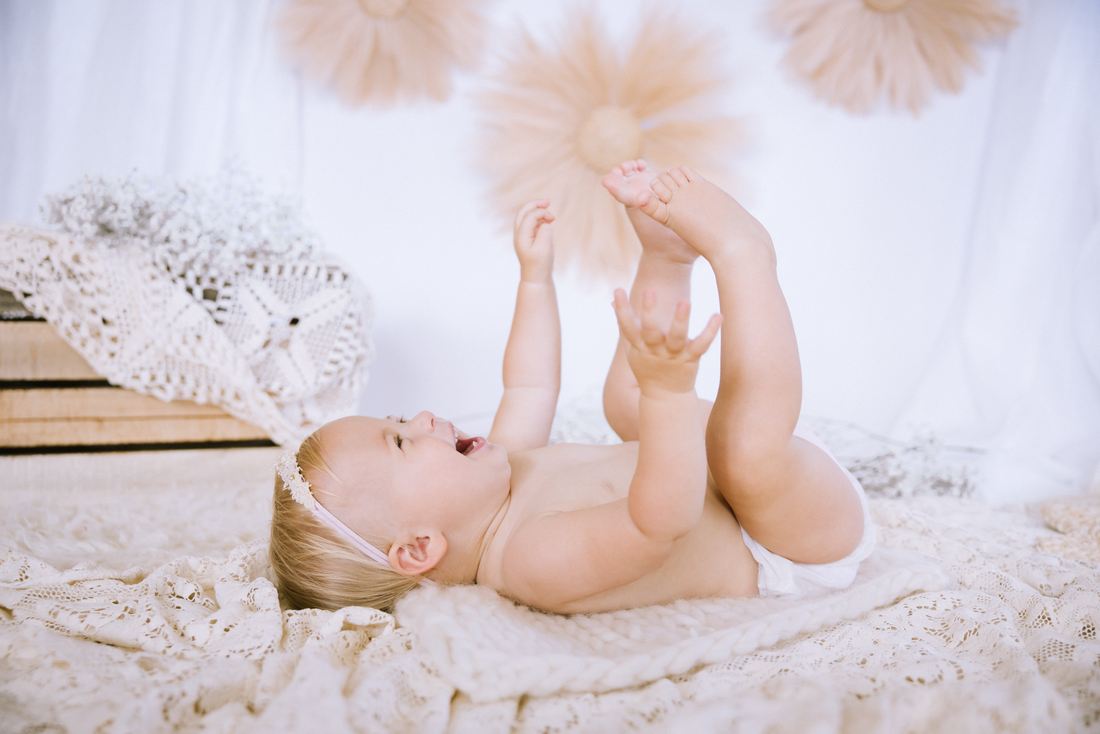A baby on her back laughing and grabbing her own toes on a lace blanket