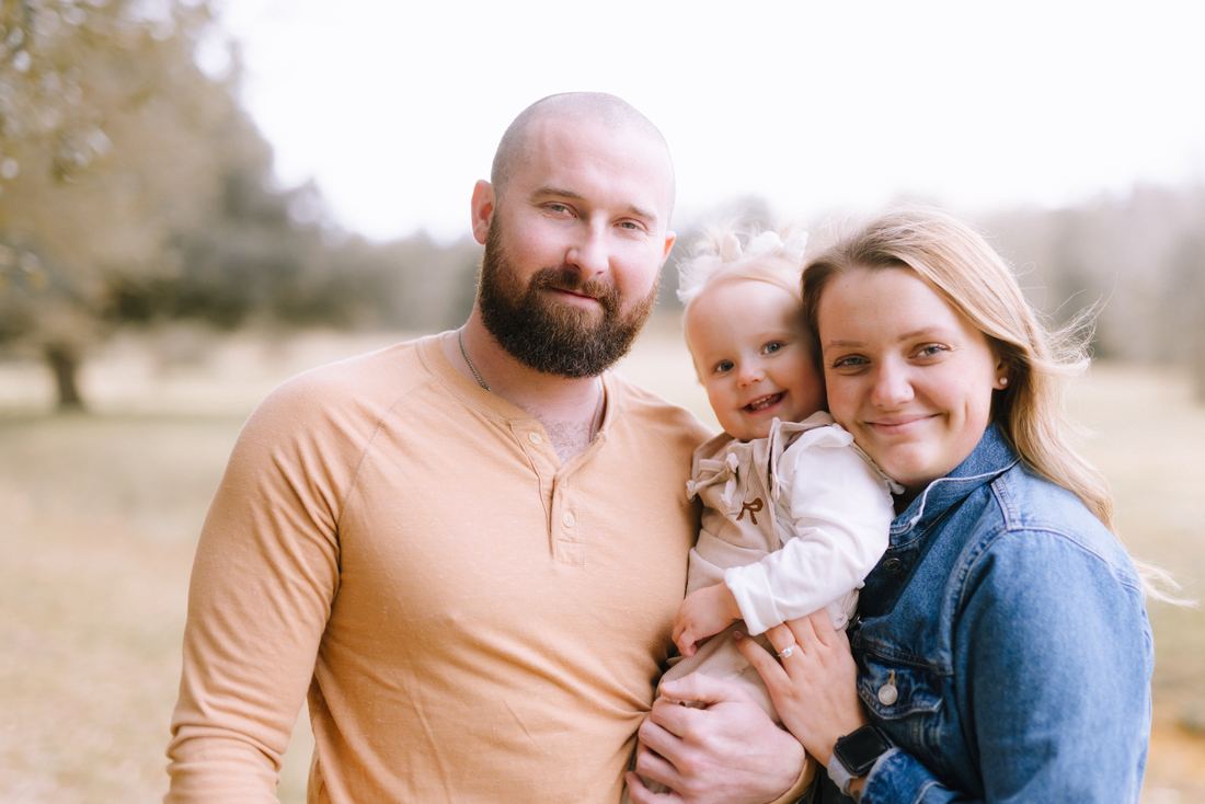 A mother and father standing close with their toddler daughter tucked between them, all three smiling at the camera in a winter field