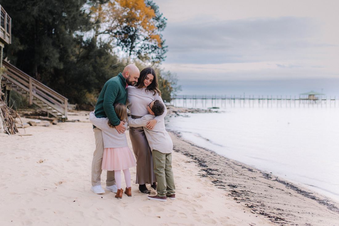 A family of four holding each other in a huddle on the sand, the bay and a long pier stretching out behind them
